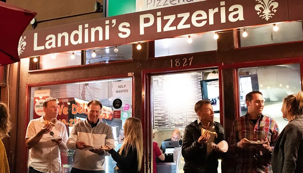 People are enjoying pizza outside Landinis Pizzeria under a warmly lit sign