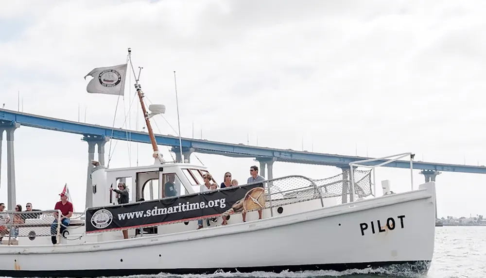 A small boat named PILOT with people aboard is sailing near a bridge displaying a banner for wwwsdmaritimeorg