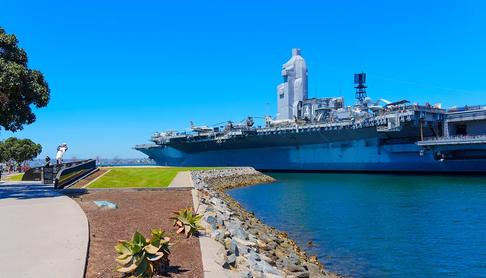 A large aircraft carrier is docked near a waterfront park with a person taking photos of it