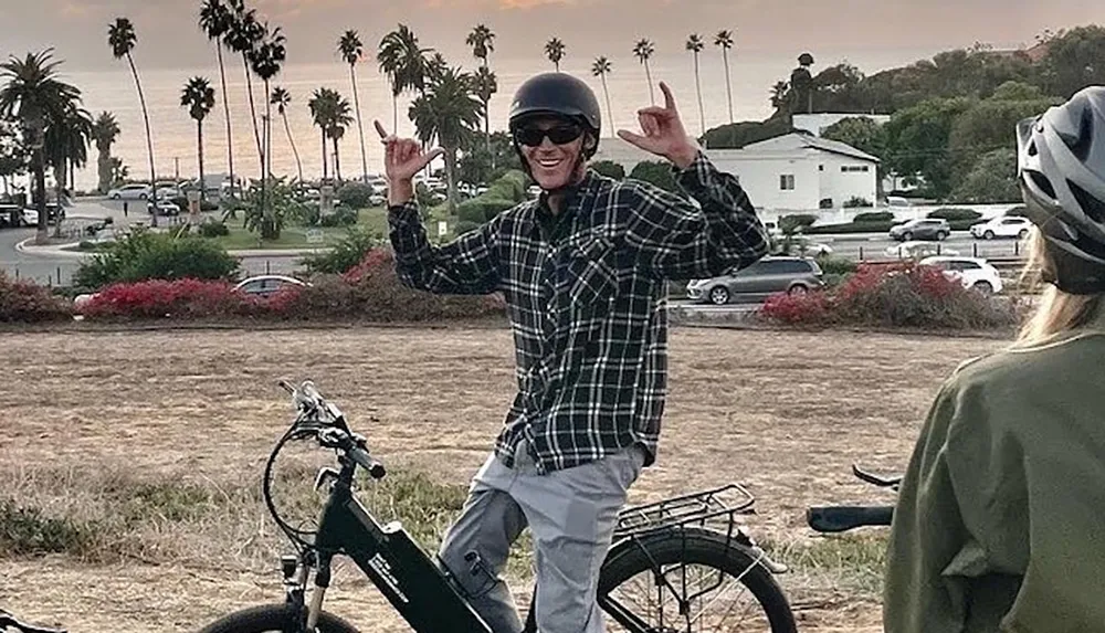 A person wearing a helmet and sunglasses is happily posing on an electric bicycle near a scenic coastal area with palm trees