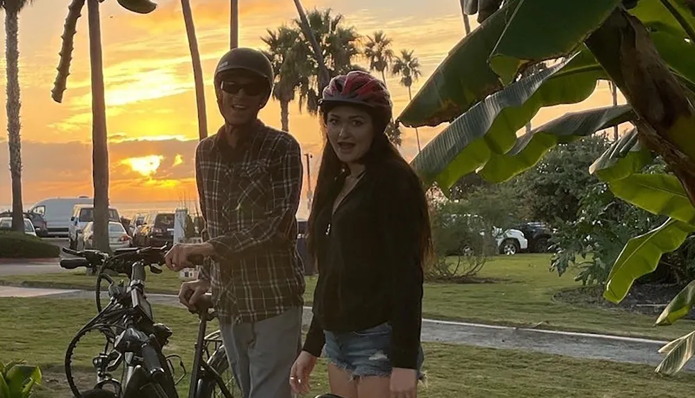 Two people wearing helmets stand with bicycles in front of a sunset backdrop with palm trees