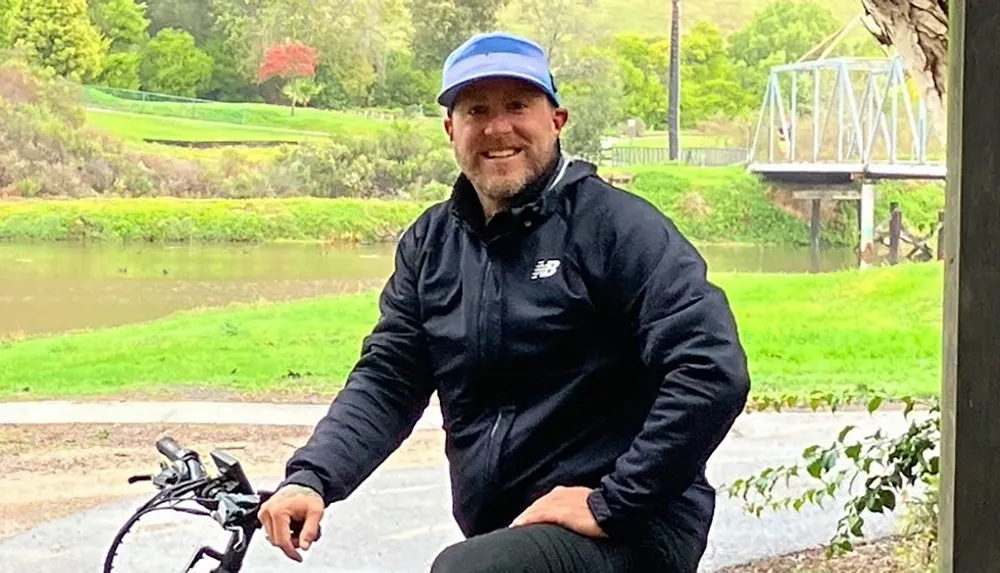 A man in a black jacket and blue cap is smiling while sitting on a bicycle in a park-like setting with a river and bridge in the background