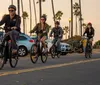 Four people are riding bicycles along a road lined with cars and palm trees at sunset