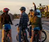 Four people are riding bicycles along a road lined with cars and palm trees at sunset