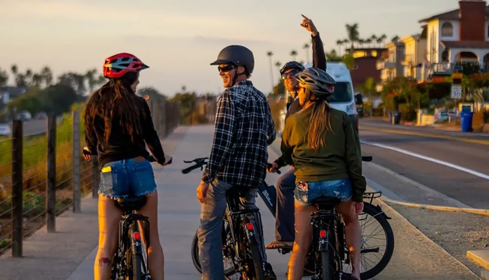 Four people wearing helmets are paused on their bicycles on a scenic path during a sunny day with one person pointing at something in the distance