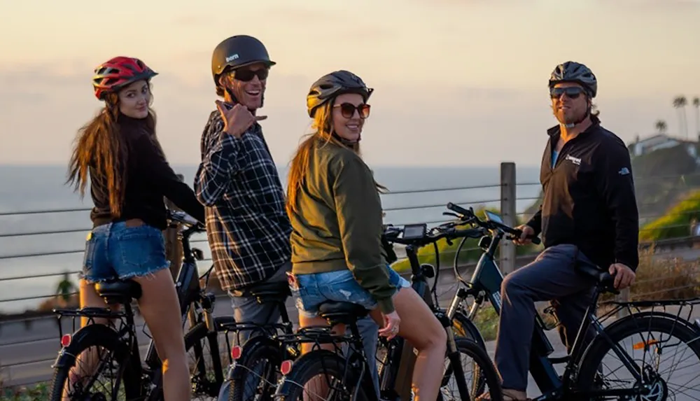 Four people wearing helmets are seated on bicycles smiling and enjoying a scenic view