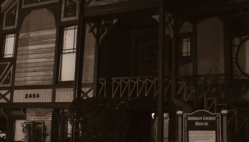 The image depicts the exterior of the Sherman Gilbert House at night showcasing its intricate architectural details and a sign with its name