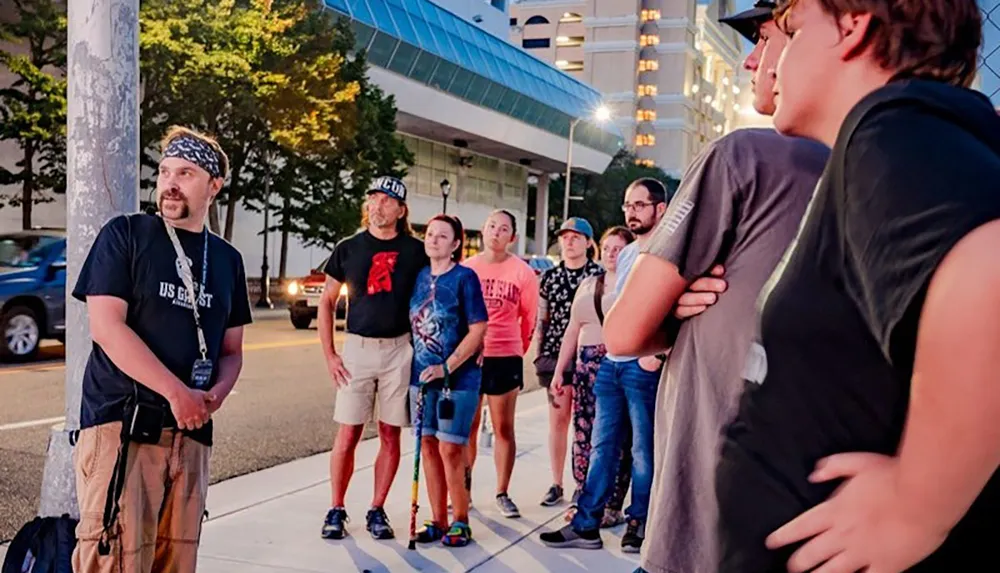 A group of people stands on a sidewalk at dusk with one person in the foreground wearing a headband and speaking