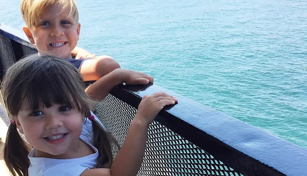 Two smiling children are standing at a railing overlooking the water