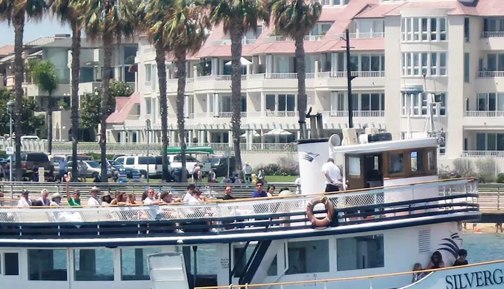 A ferry boat with passengers is docked near a waterfront lined with palm trees and residential buildings