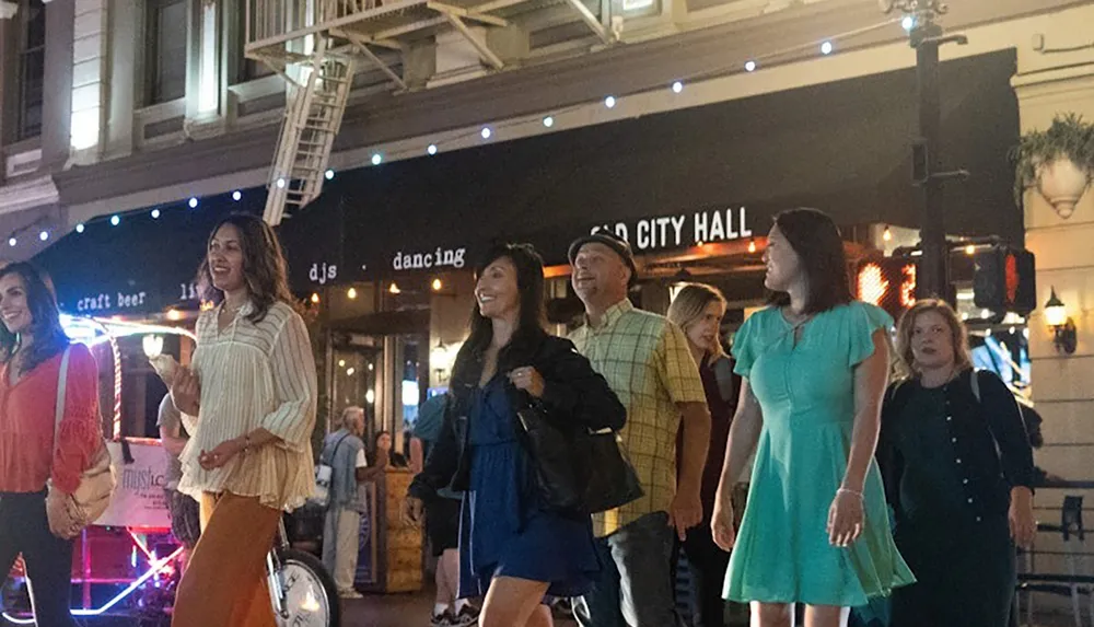 A group of people are happily walking down a lively street at night past a venue with signs for craft beer live DJs and dancing