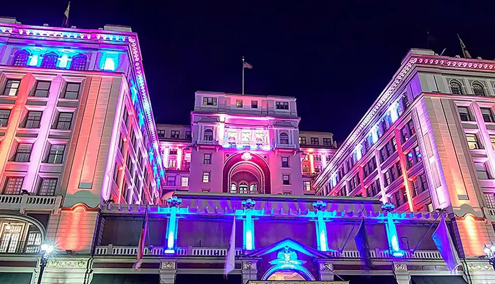 The image shows a grand building illuminated with vibrant purple and blue lights against a night sky