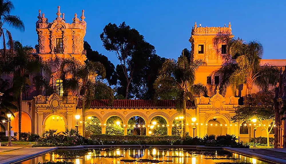 The image showcases an ornate Spanish-style building with arches and towers surrounded by palm trees reflecting in a tranquil pond at dusk