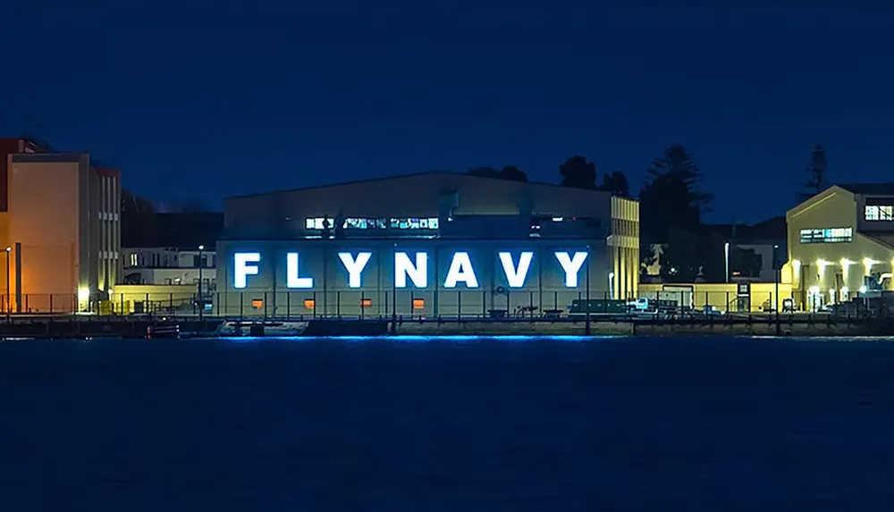 The image shows a building at night with large illuminated letters spelling FLY NAVY