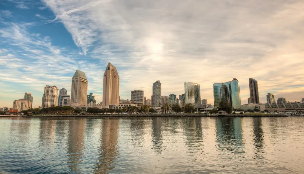 A city skyline is reflected in the calm waters under a sky with scattered clouds