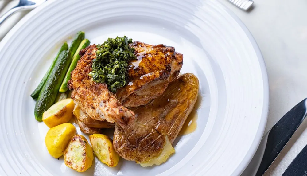 A dish featuring grilled chicken topped with a green herb sauce served with roasted potatoes and green vegetables on a white plate