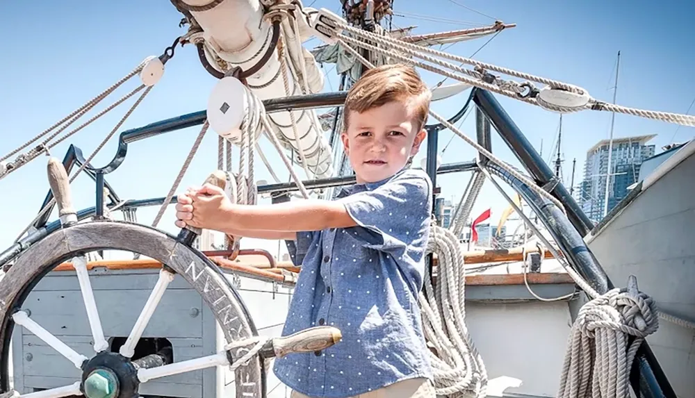 A young boy is steering a ships wheel on a sailboat under a clear blue sky