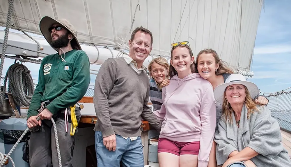 A group of people pose happily on a sailboat under clear skies with large sails visible in the background