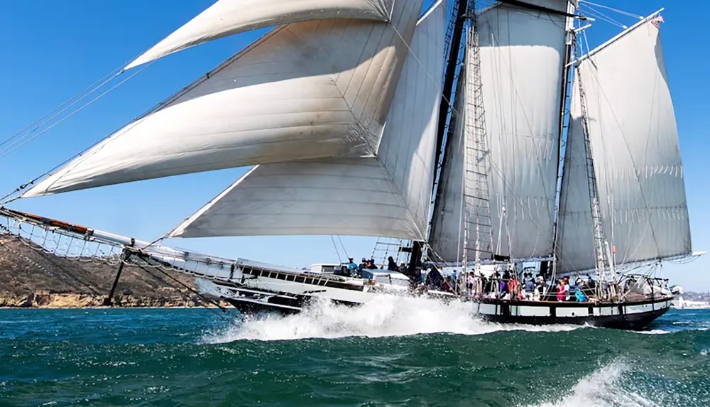 A large sailing ship with white sails is cutting through the ocean waves under a clear blue sky