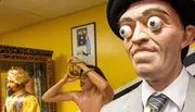 The image shows a wax figure with exaggerated bulging eyes in a museum setting, featuring other intriguing exhibits in the background.