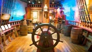 The image depicts a colorful, interactive pirate ship exhibit with a ship's wheel, barrels, and nautical decorations.