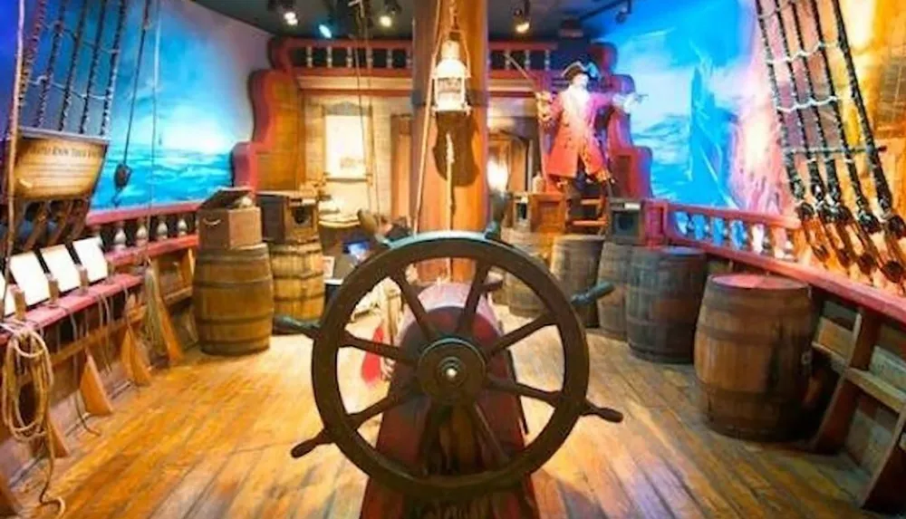 The image depicts a colorful interactive pirate ship exhibit with a ships wheel barrels and nautical decorations