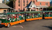 A group of people is riding a tour trolley in a town with historic-looking buildings in the background.