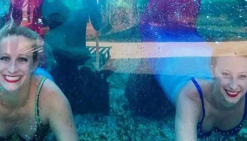 Two smiling women dressed as mermaids are underwater posing behind a glass panel