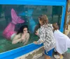 Two children are looking at a woman dressed as a mermaid inside an aquarium display