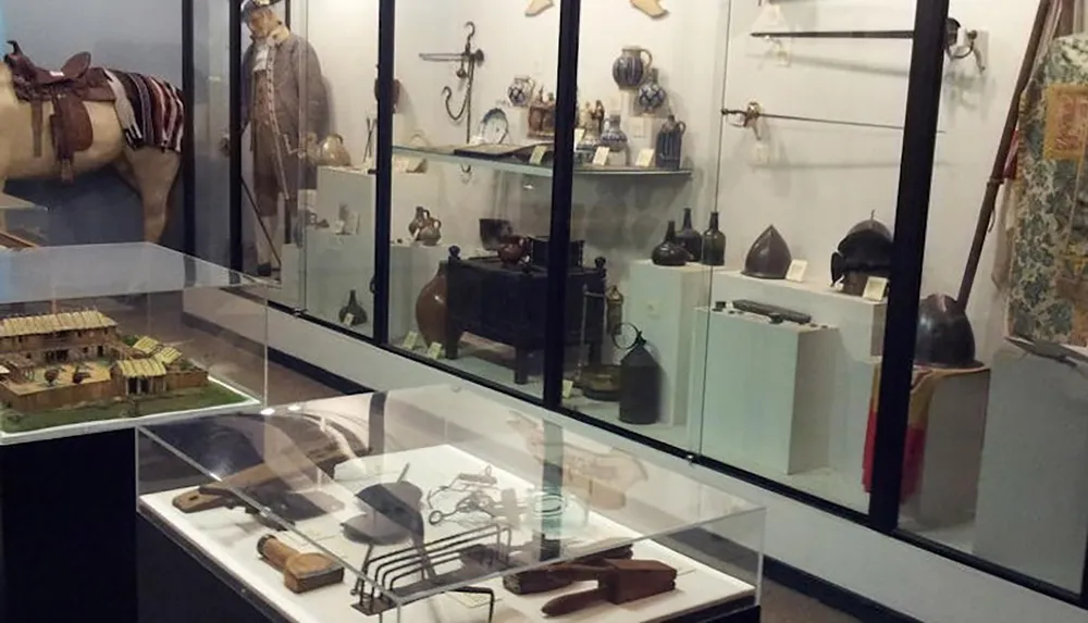 The image shows a museum exhibit featuring historical artifacts including old tools pottery helmets and a model of a building displayed in glass cases