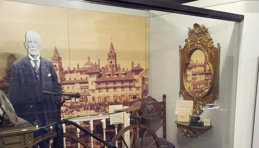 The image shows a museum exhibit featuring a historic photo of a man next to a vintage bicycle and ornate framed decor with a backdrop of an old architectural building