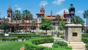 The image shows a formal garden with manicured hedges and a statue in the foreground, set against the backdrop of a historic building with red-tiled roofs and towers.