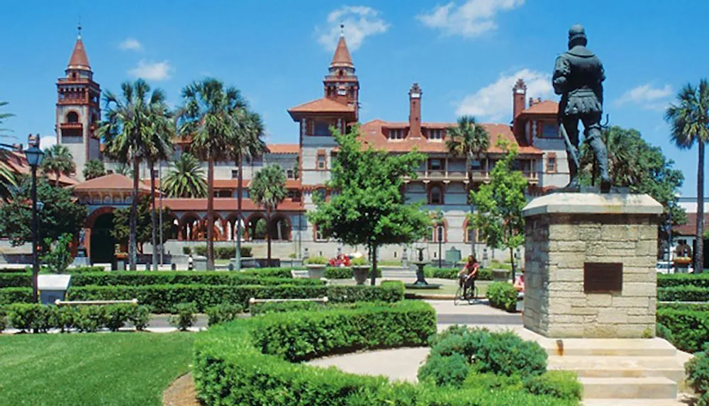 The image shows a formal garden with manicured hedges and a statue in the foreground set against the backdrop of a historic building with red-tiled roofs and towers