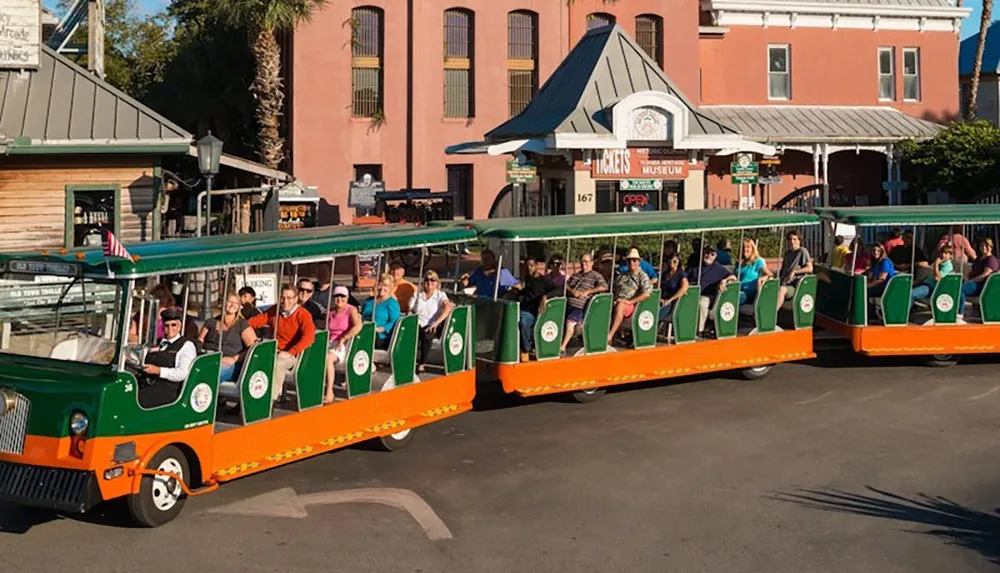 A group of people are seated in an open-air trolley on a sunny day in front of a building with a sign that reads Tickets and Museum