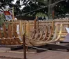 The image shows a partially constructed wooden ship frame in an outdoor setting surrounded by fencing and flags