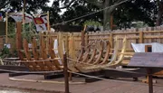 The image shows a partially constructed wooden ship frame in an outdoor setting, surrounded by fencing and flags.