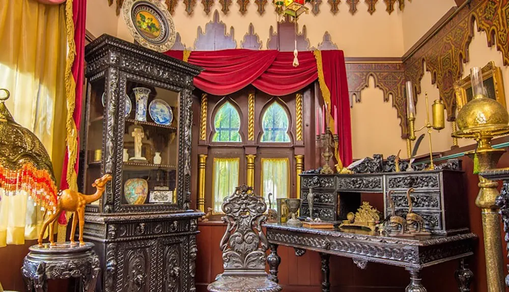 The image showcases an ornate room filled with intricately carved dark wood furniture decorative artifacts and vibrant red drapery accenting the arched windows