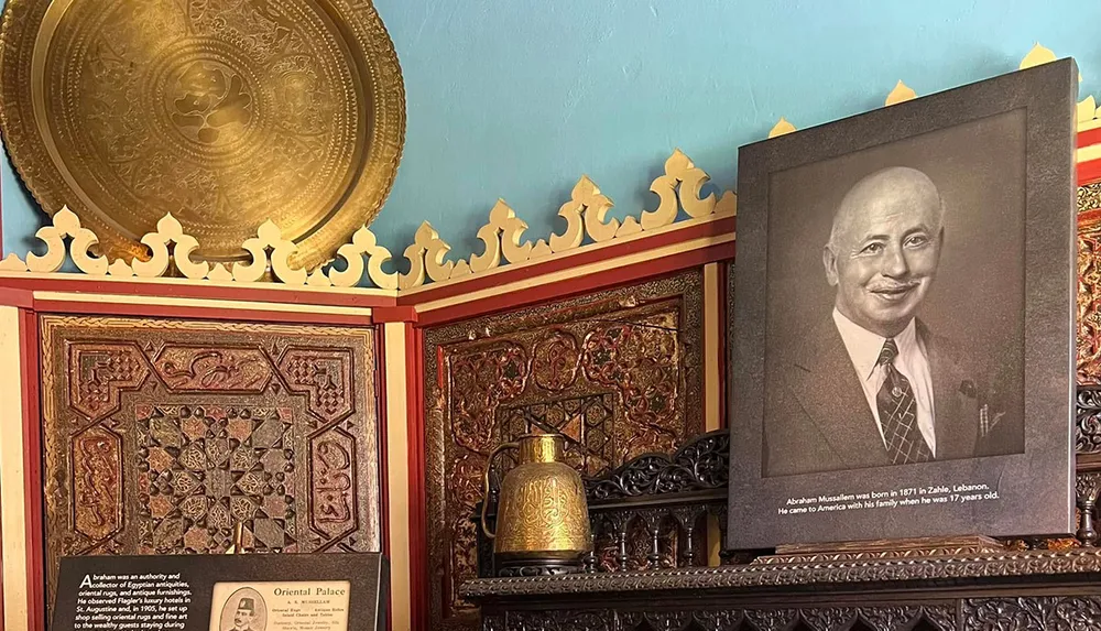 The image shows a room adorned with ornate intricate decorations featuring a large circular brass plate carved wooden panels and a prominently displayed portrait of a man with a description about him underneath