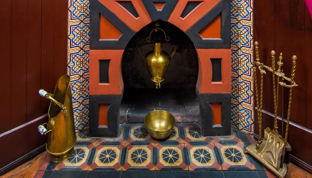 The image features a decorative fireplace with colorful geometric tile patterns a brass kettle hanging inside and brass fireplace tools nearby