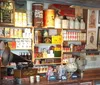 The image shows a vintage-style general store interior filled with colorful tins a large red cast iron grinder and various old-fashioned goods displayed on shelves and a counter