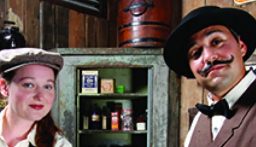 Two people dressed in vintage attire stand smiling in front of an old cabinet filled with various items