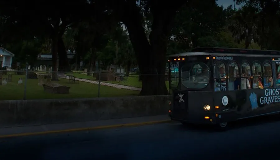 A black tour bus labeled Ghosts & Gravestones passes by a cemetery at night.