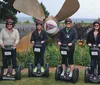 Four people wearing helmets are standing in a line on Segways in front of a large airplane propeller sculpture