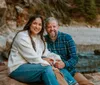 A smiling woman and man are sitting closely together on a rock by a riverbank surrounded by a natural wooded environment