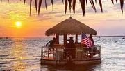 A tiki bar-style floating deck with people on board flutters the American flag against a backdrop of a sunset over serene waters.