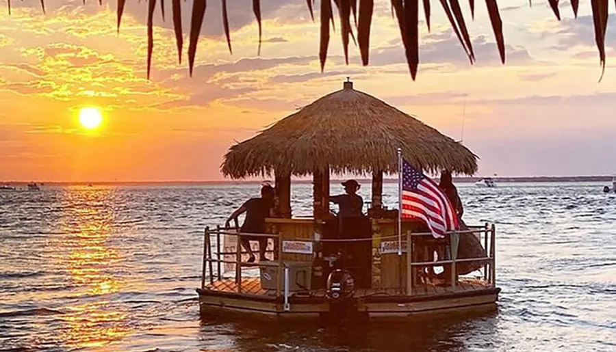 A tiki bar-style floating deck with people on board flutters the American flag against a backdrop of a sunset over serene waters.