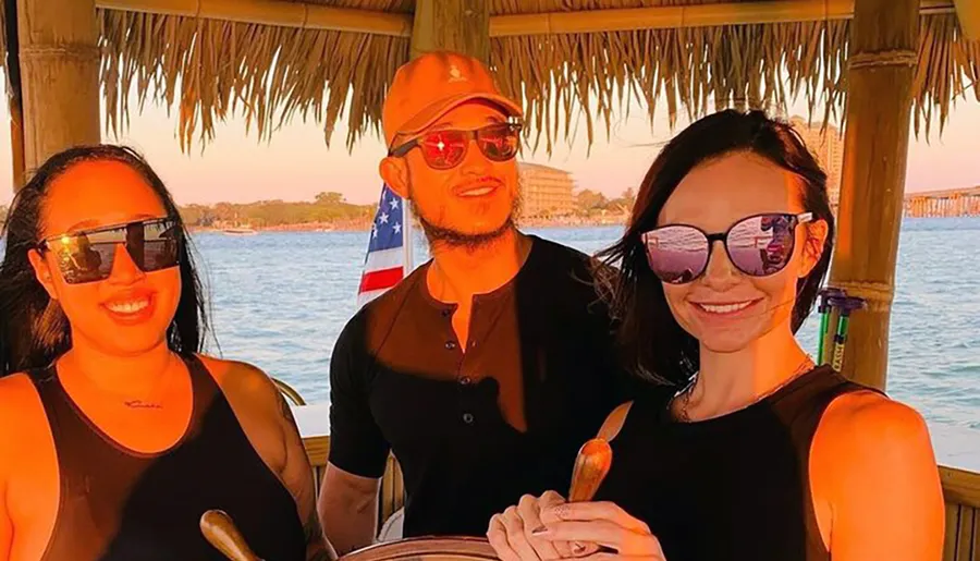 Three people are smiling and posing together under a thatched roof on a boat with a backdrop of water and a flag at sunset.