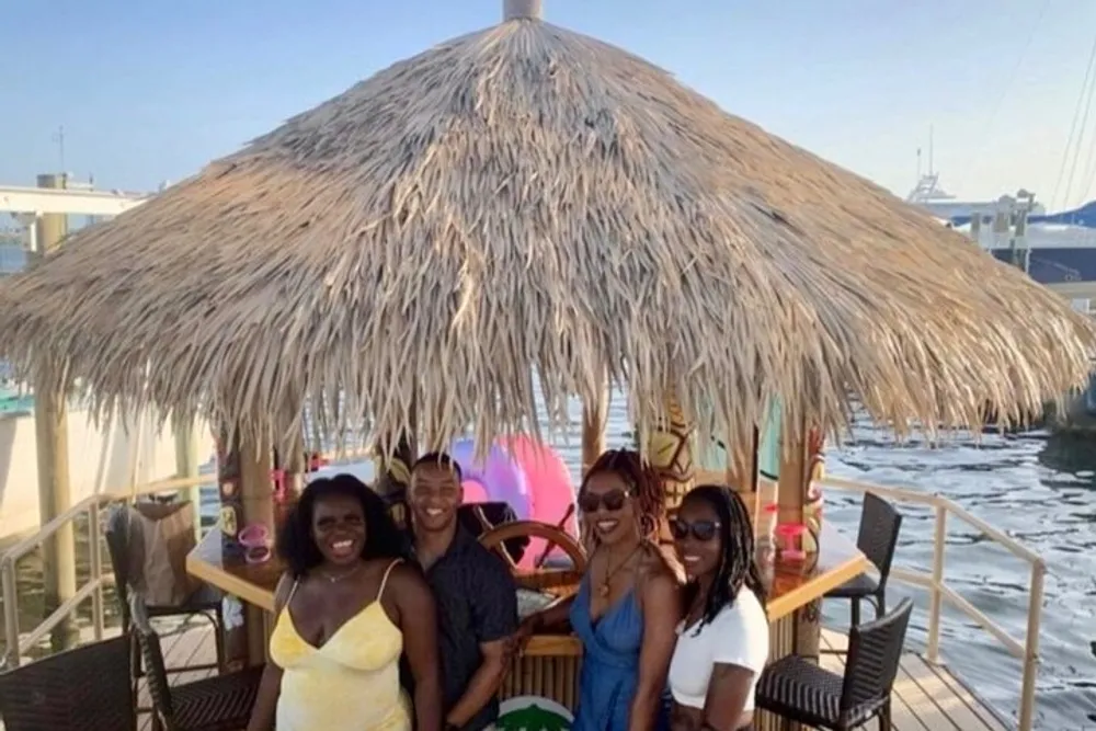 Four people are smiling in front of a thatched-roof structure by the water possibly enjoying a social outing or vacation