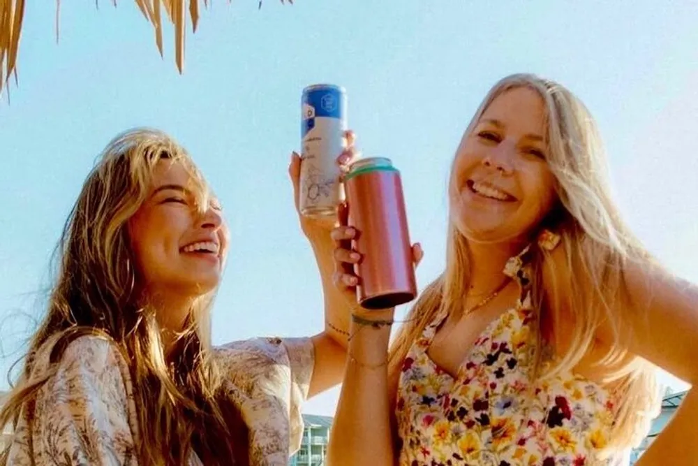 Two individuals are smiling and toasting with a can and a tumbler against a sunny outdoor background