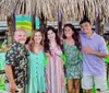 Five people are smiling for a group photo at a tropical-themed outdoor venue with tiki-style decorations and a water slide visible in the background